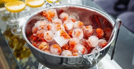 A stainless steel bowl has various-shaped ice cubes infused with fruit. Cocktail glasses are next to the bowl.