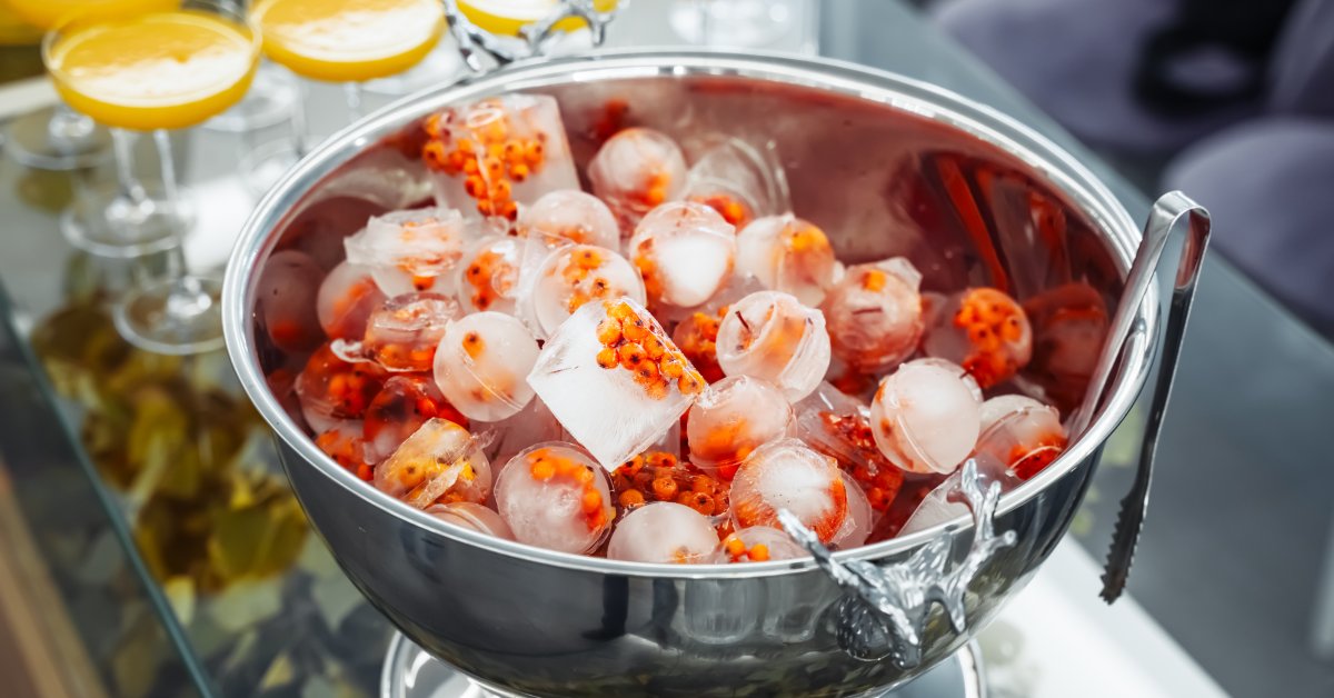 A stainless steel bowl has various-shaped ice cubes infused with fruit. Cocktail glasses are next to the bowl.