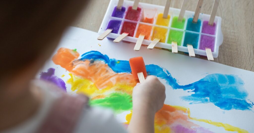 An ice cube tray with various paint-infused ice. A child uses a red ice cube to paint on a piece of paper.