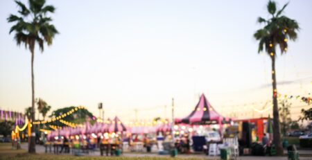 A blurred fair with pink-and-black tents set up on a street. There are twinkle lights and palm trees decorating the scene.