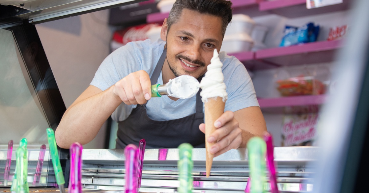 A man stands inside an ice cream truck. He smiles as he scoops vanilla ice cream into a cone that he holds in his other hand.