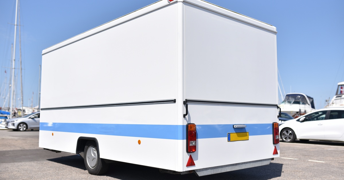 A white trailer with a blue stripe is parked in a parking spot. Other cars and boats are in the background.