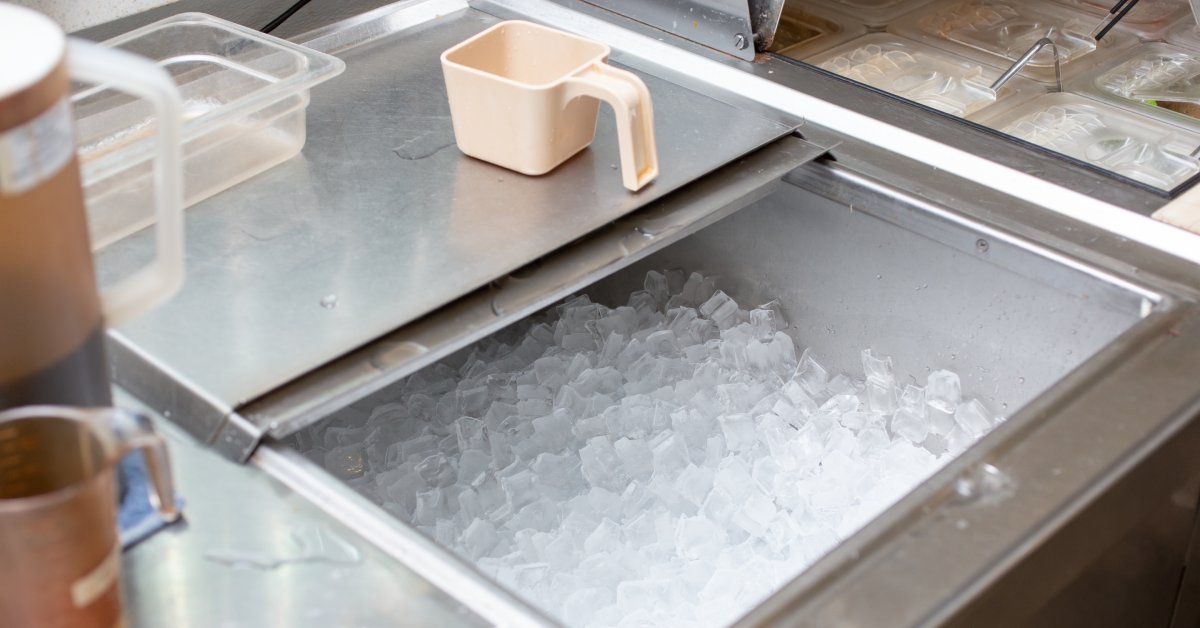 A cooler of ice inside a restaurant. The top lid is open with a condiment section to the right of the cooler.