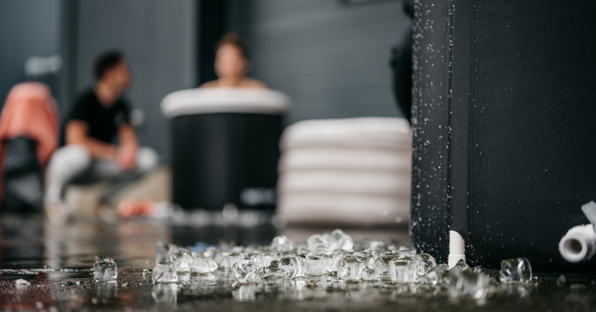 A close-up of a pile of ice on the floor next to an ice bath. Someone is sitting in a bath in the background.