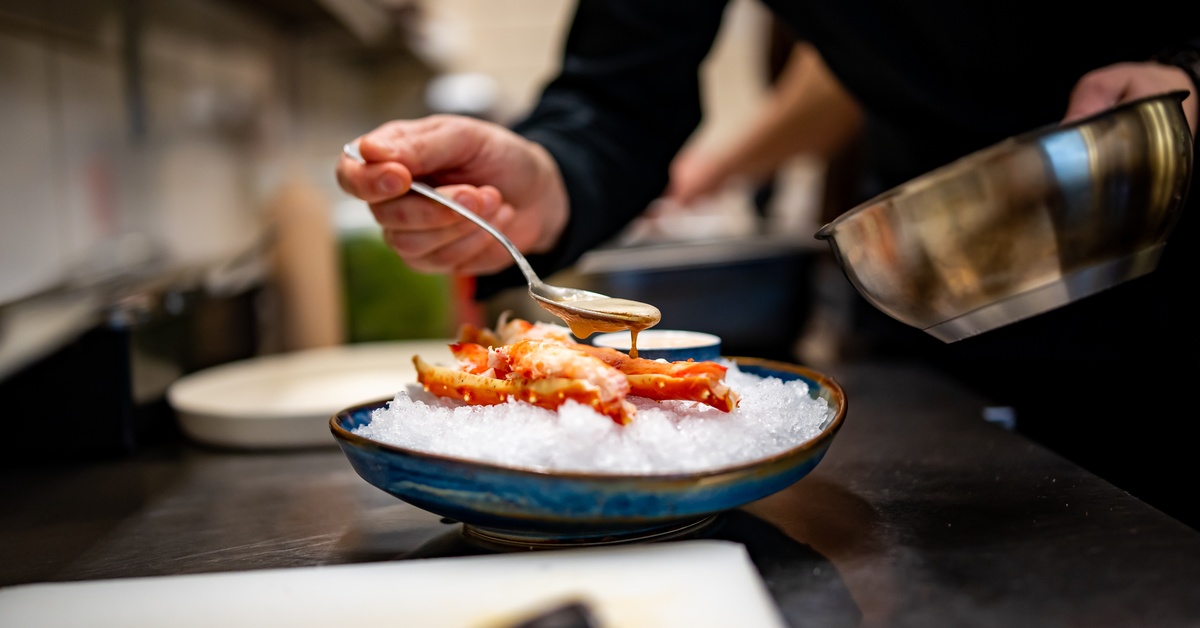 A close-up of crab legs resting on top of a bowl of ice. A chef drizzles a sauce on top with a spoon.