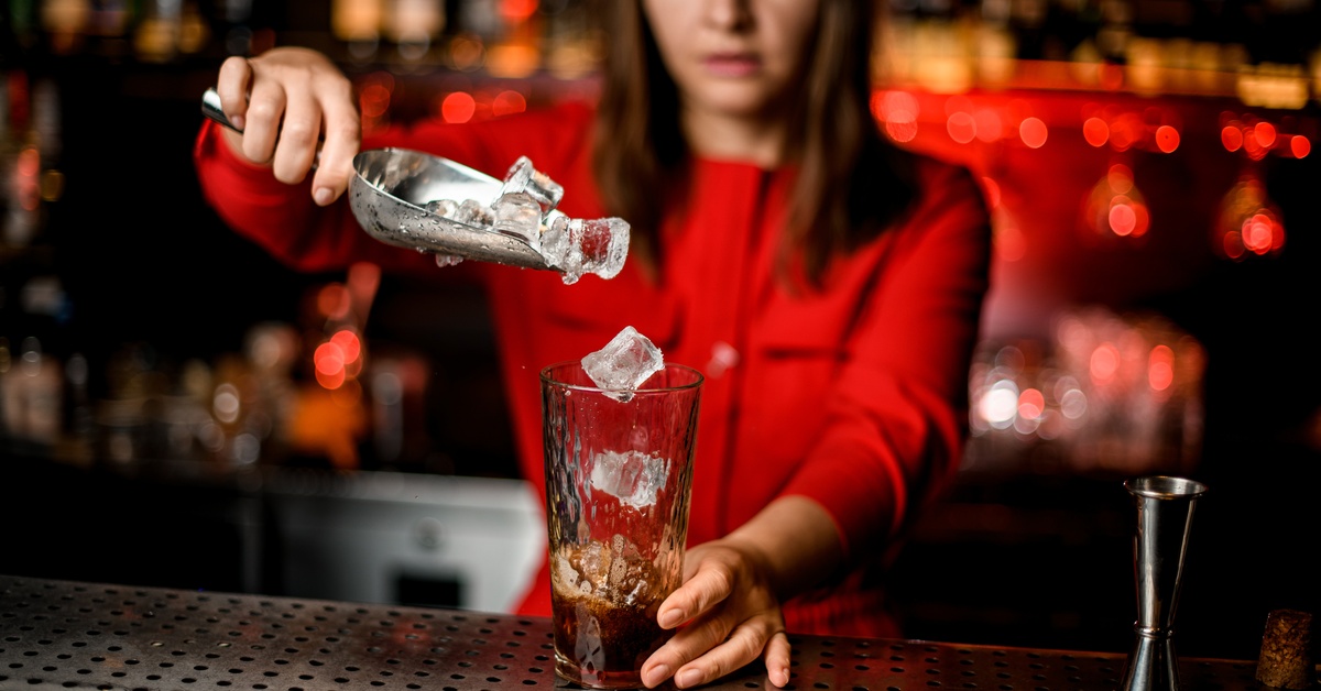 A woman stands behind a bar with a glass in front of her. She uses a metal scoop to pour ice into a glass.