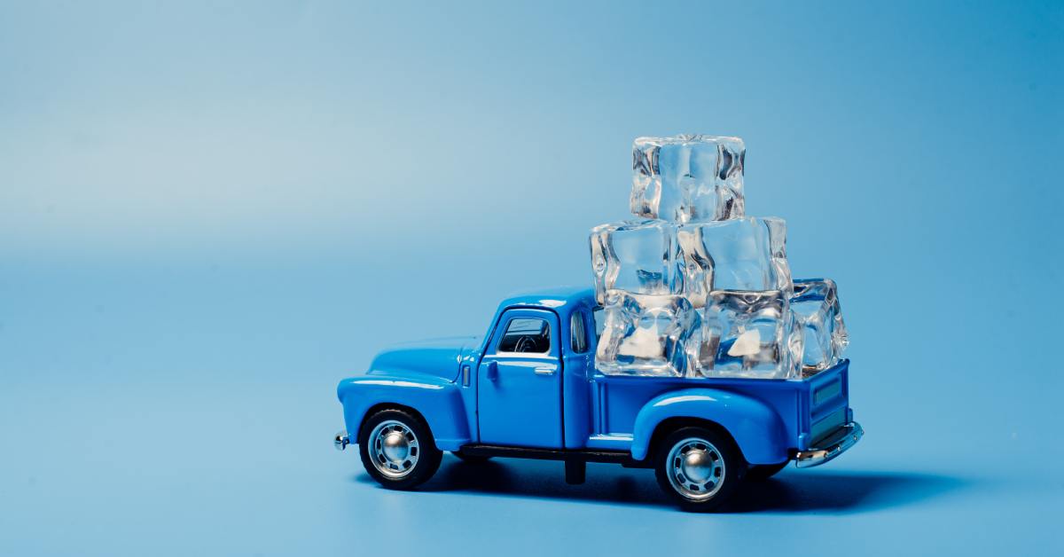 A small, blue toy truck carrying a stack of clear ice cubes in the back. The truck and ice rest against a light-blue background.