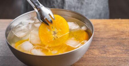 A metal bowl filled with water, ice cubes, and orange slices. Someone uses a pair of tongs to pull out one of the slices.