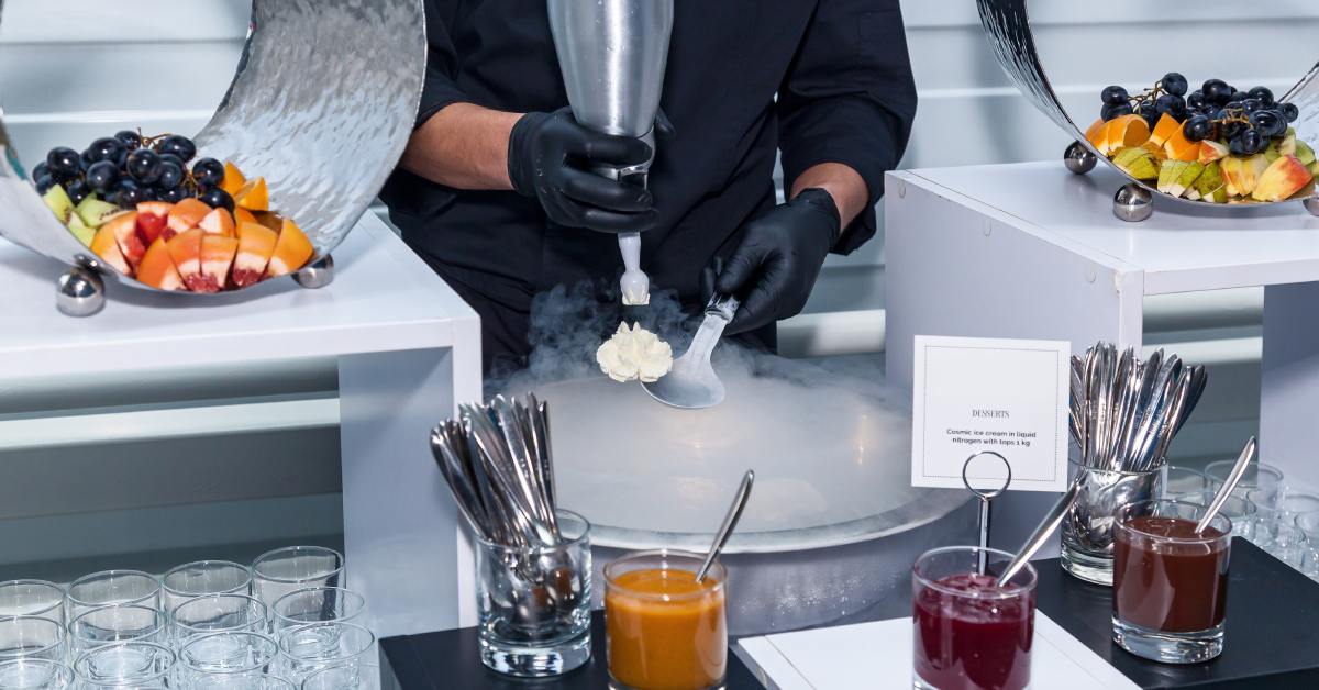 A gloved chef pipes cream onto a spoon over a foggy, chilled pan. Fruit platters, sauces, and glassware surround the chef.