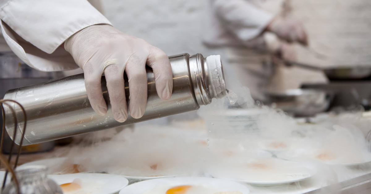 A gloved hand pours liquid nitrogen from a metal canister, creating a rolling white fog over small plates on a table.
