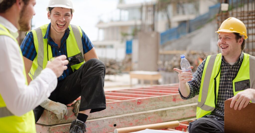 Three construction workers wearing PPE sit and stand in a circle. They laugh while one holds a water bottle.