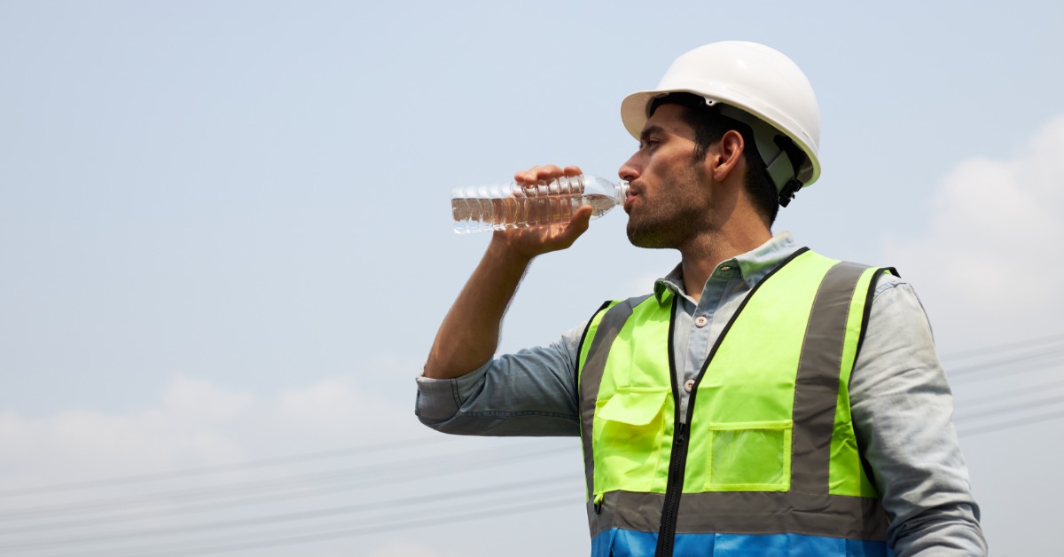 A construction worker wearing a safety vest and a hard hat drinks from a clear plastic water bottle.