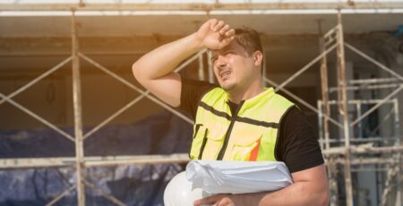 A construction worker wearing a safety vest shades his eyes from the sun with his hand. He holds a hard hat in his other hand.