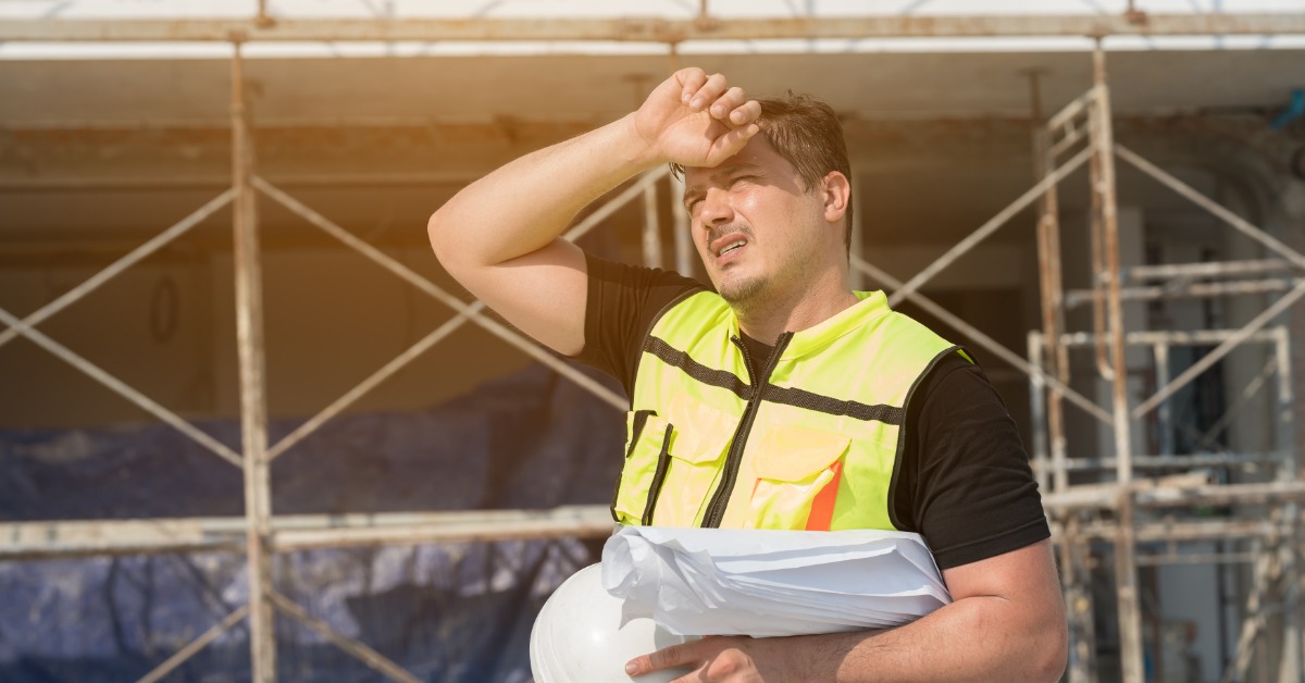 A construction worker wearing a safety vest shades his eyes from the sun with his hand. He holds a hard hat in his other hand.