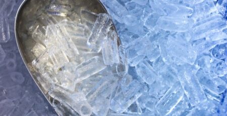A close-up of a metal scoop immersed in a pile of ice cubes. The ice cubes are cylindrical with a hole in the middle.
