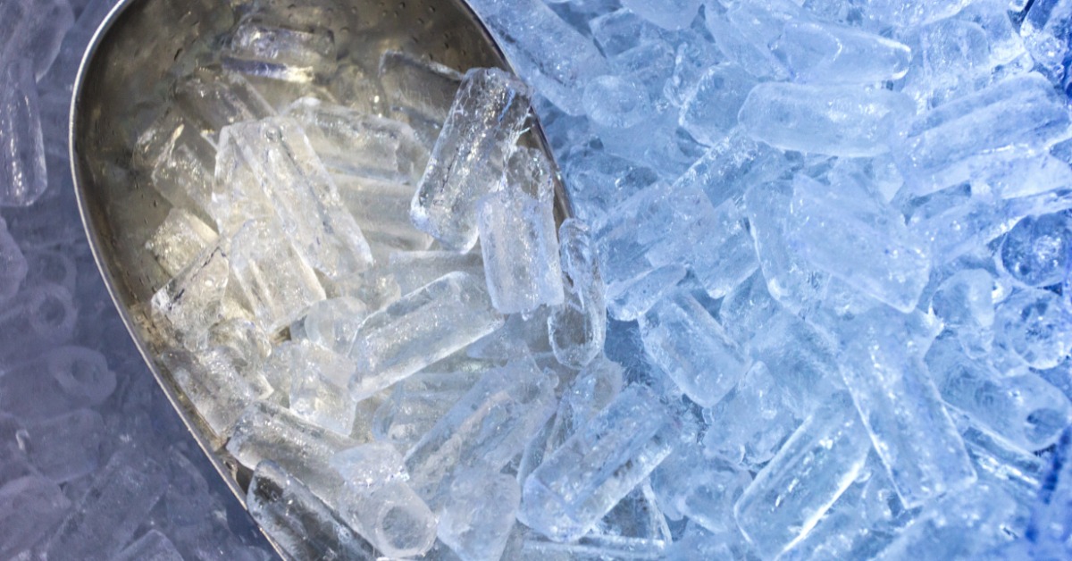 A close-up of a metal scoop immersed in a pile of ice cubes. The ice cubes are cylindrical with a hole in the middle.