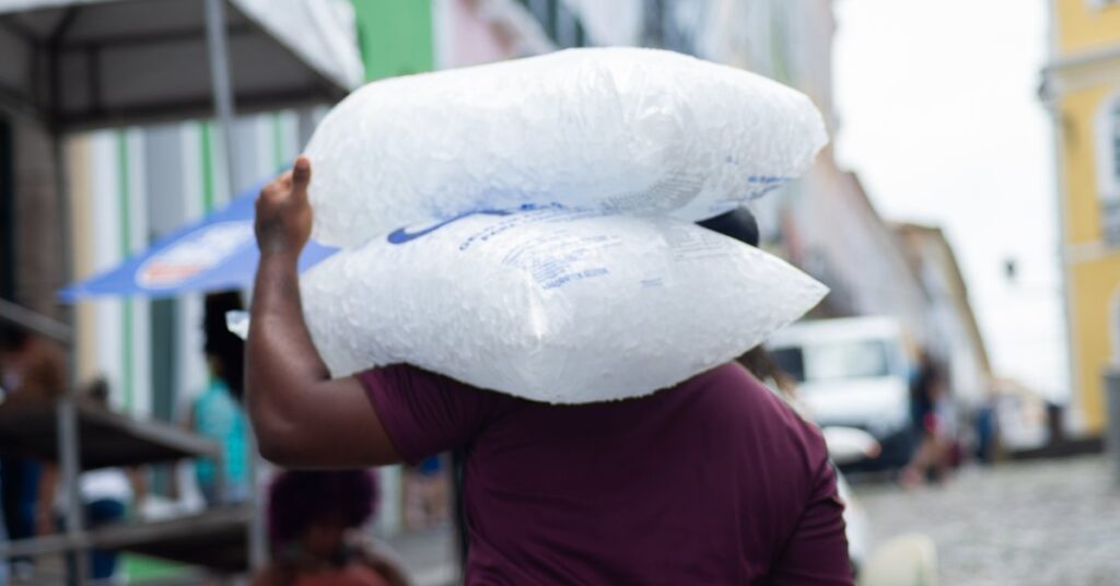 Someone carries two very large bags of ice over their shoulder. People and cars are blurred on the street ahead.