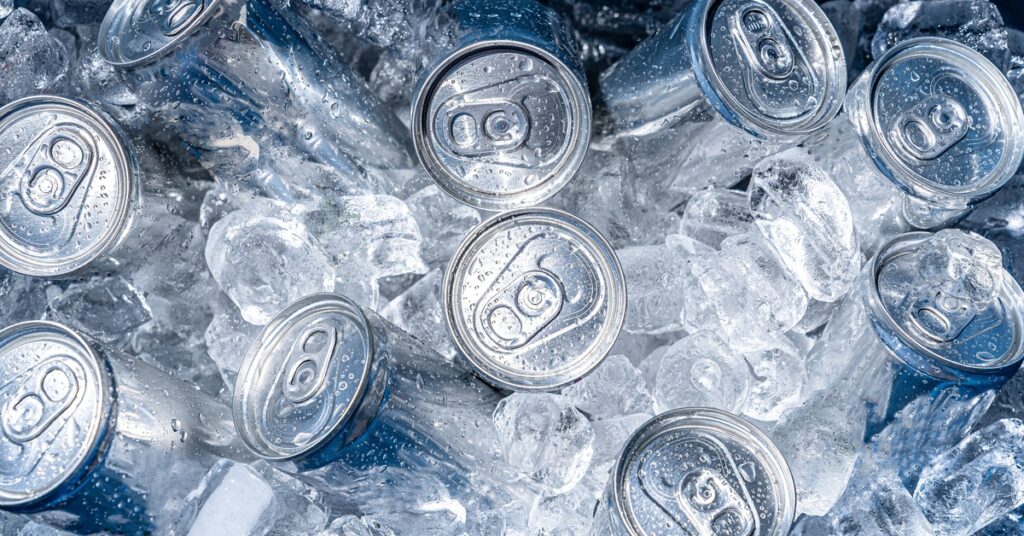 A close-up of the inside of a cooler. The tops of several cans peak out over a mound of ice cubes covering the drinks.
