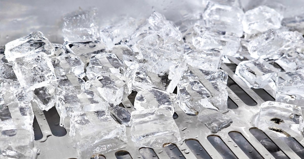 A pile of clear ice cubes melts across a slotted metal surface, leaving droplets and small puddles between the cubes.