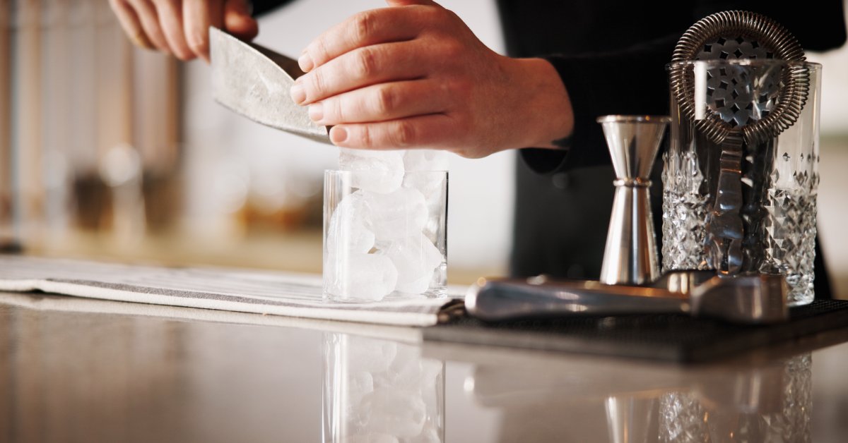 A bartender scoops crushed ice into a glass behind the bar as cocktail tools and another glass sit nearby.