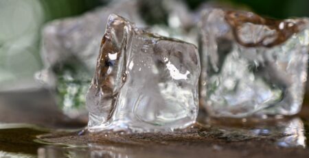 Several clear ice cubes melt on a wet surface as warm outdoor air softens their edges and forms small puddles.