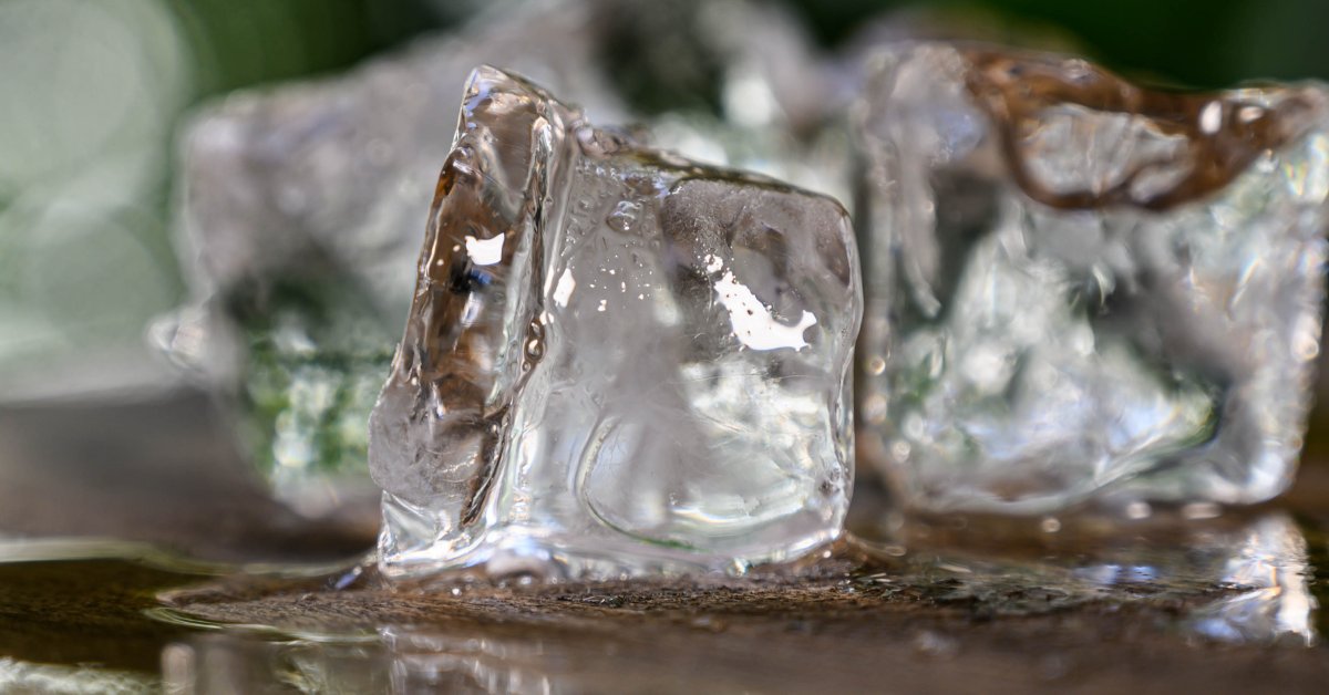 Several clear ice cubes melt on a wet surface as warm outdoor air softens their edges and forms small puddles.