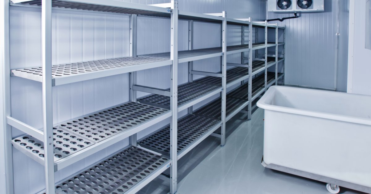 An empty commercial freezer contains rows of metal shelves beside a large rolling storage bin under bright lights.