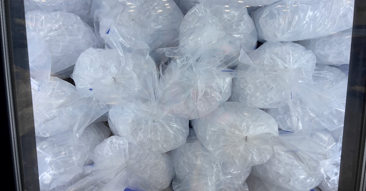 Clear plastic bags filled with ice cubes are stacked behind dark chiller doors with bright overhead reflections.