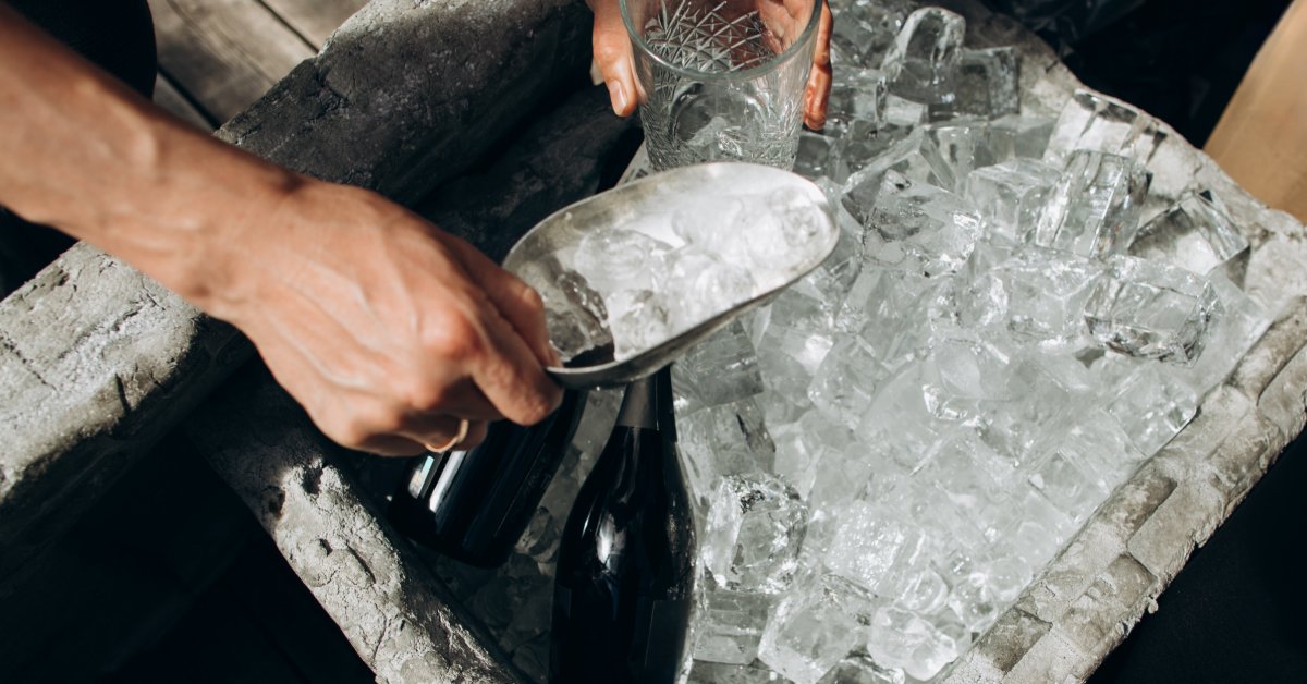 Hands using a metal scoop near a clear glass above a bin of ice cubes with dark bottles and empty glasses.